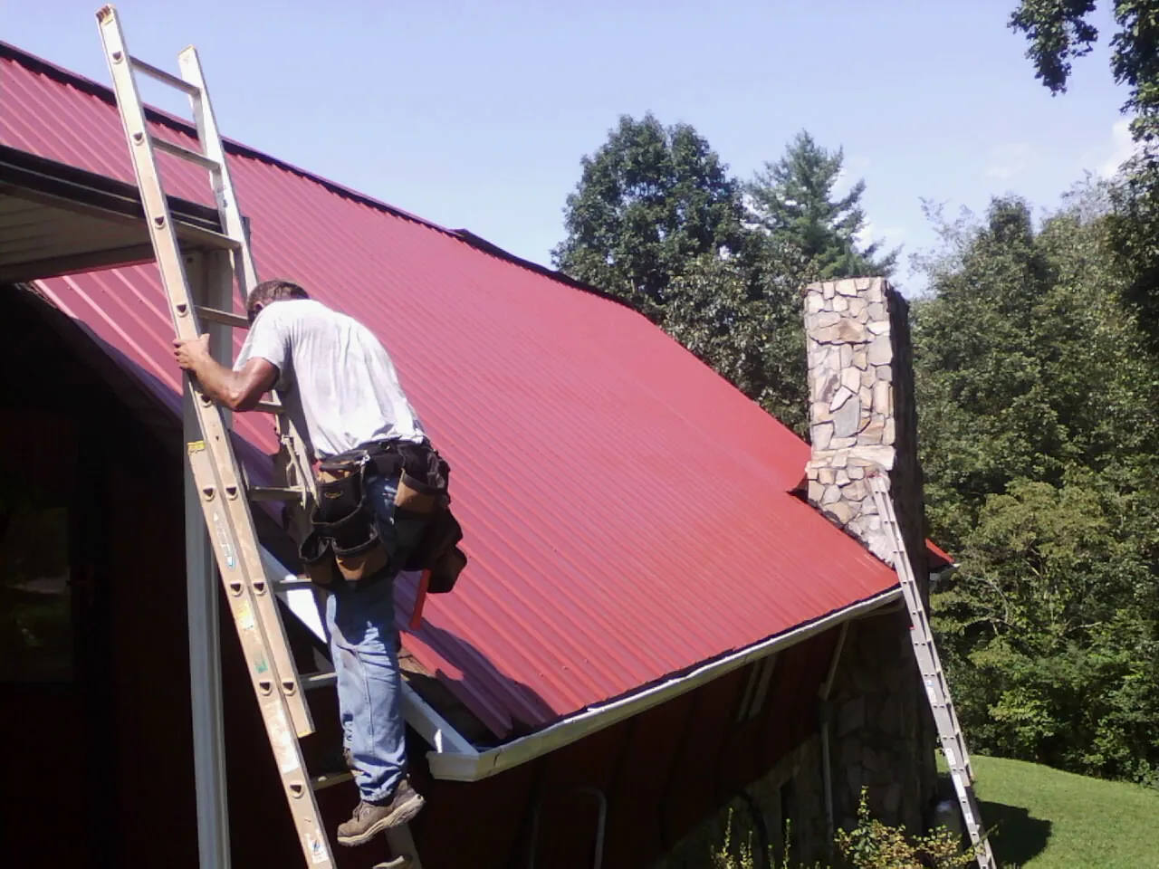 Roofer on a ladder beside a red metal roof and stone chimney — Soffit & Fascia Repair by Roofing Brothers in Perry